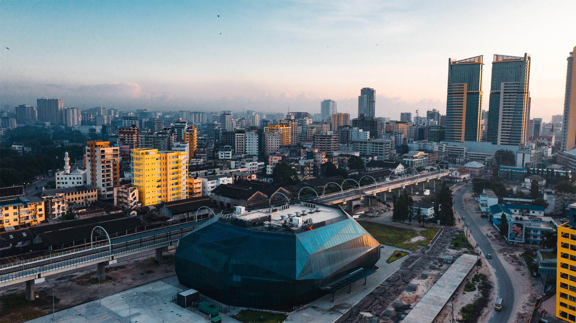A cityscape at dawn or dusk, with a gradient sky from blue to orange. Various buildings of different heights are visible, including a prominent geometric structure with a glass facade in the foreground. A train track runs through the middle, leading towards taller buildings in the background