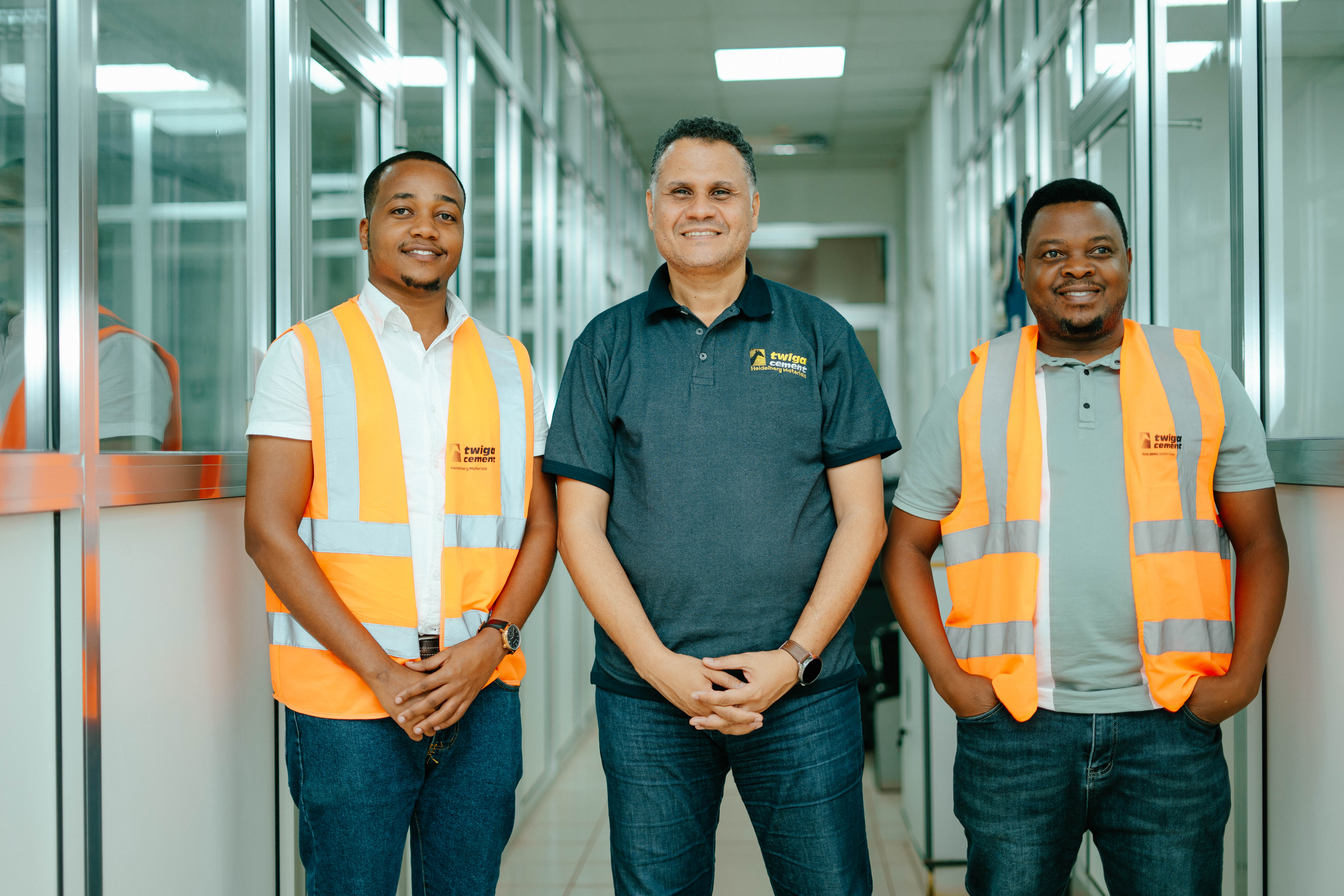 Three workers in a bright office hallway, two wearing orange safety vests and one in a branded polo shirt.