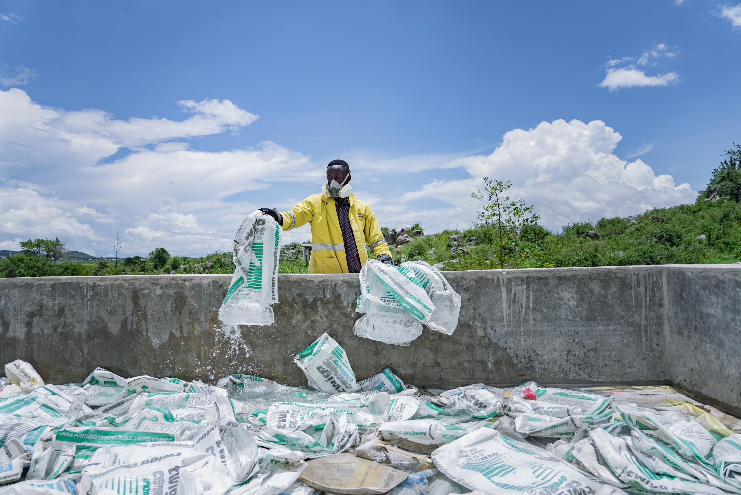 A worker wearing protective gear handles empty cement bags at an industrial site under a bright blue sky.