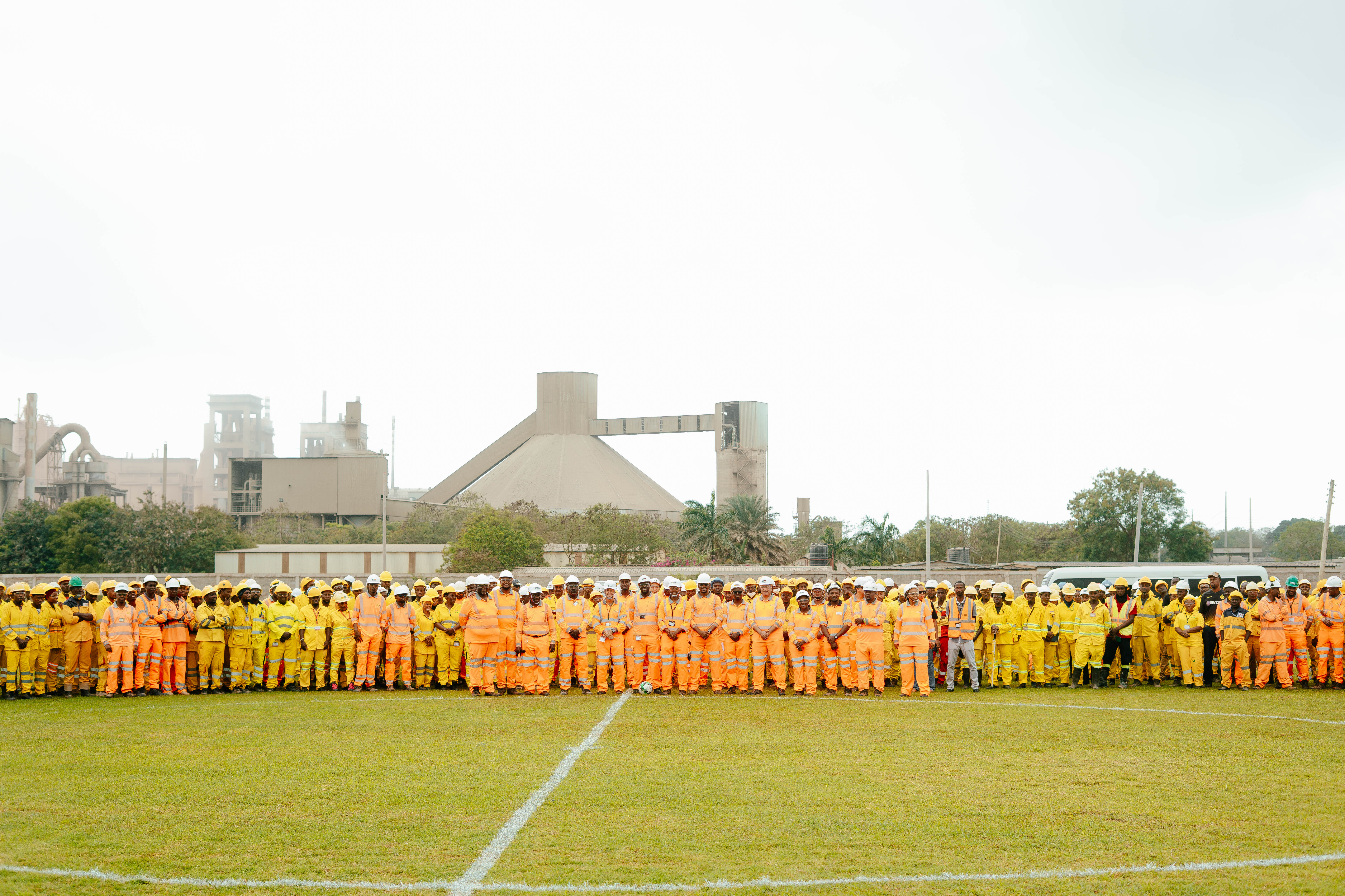 A large group of workers in yellow and orange safety uniforms stands on a grass field in front of an industrial building.
