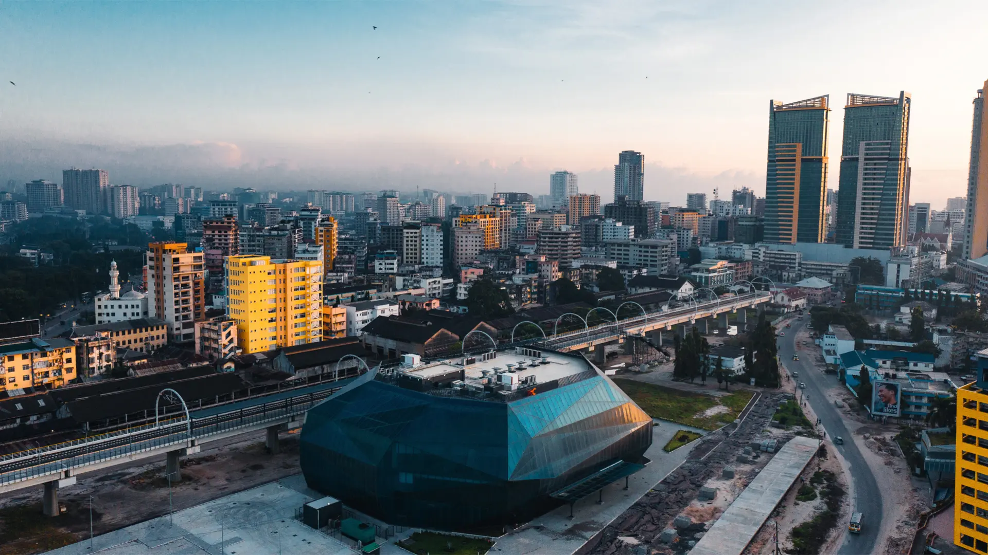 A cityscape at dawn or dusk, with a gradient sky from blue to orange. Various buildings of different heights are visible, including a prominent geometric structure with a glass facade in the foreground. A train track runs through the middle, leading towards taller buildings in the background