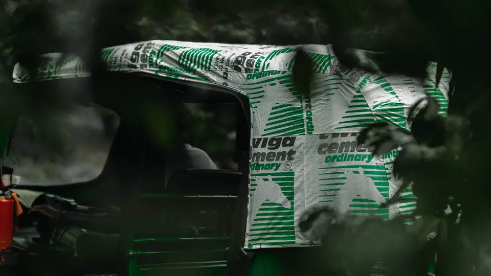 A green auto rickshaw with a roof made from Heidelberg Materials cement bags, partially obscured by foliage.